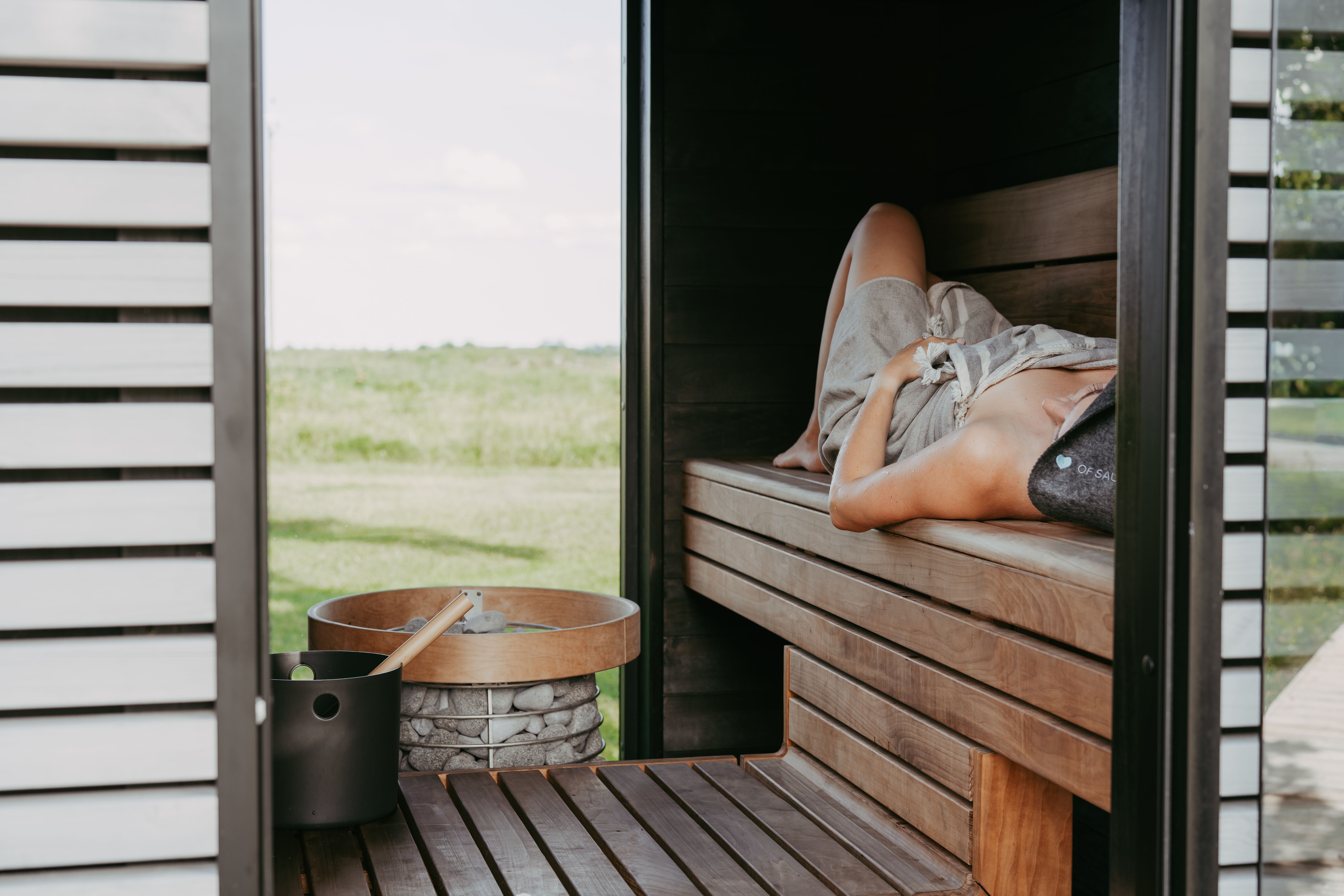 A person resting on a wooden sauna bench beside the heater, with a green field visible through the open door.