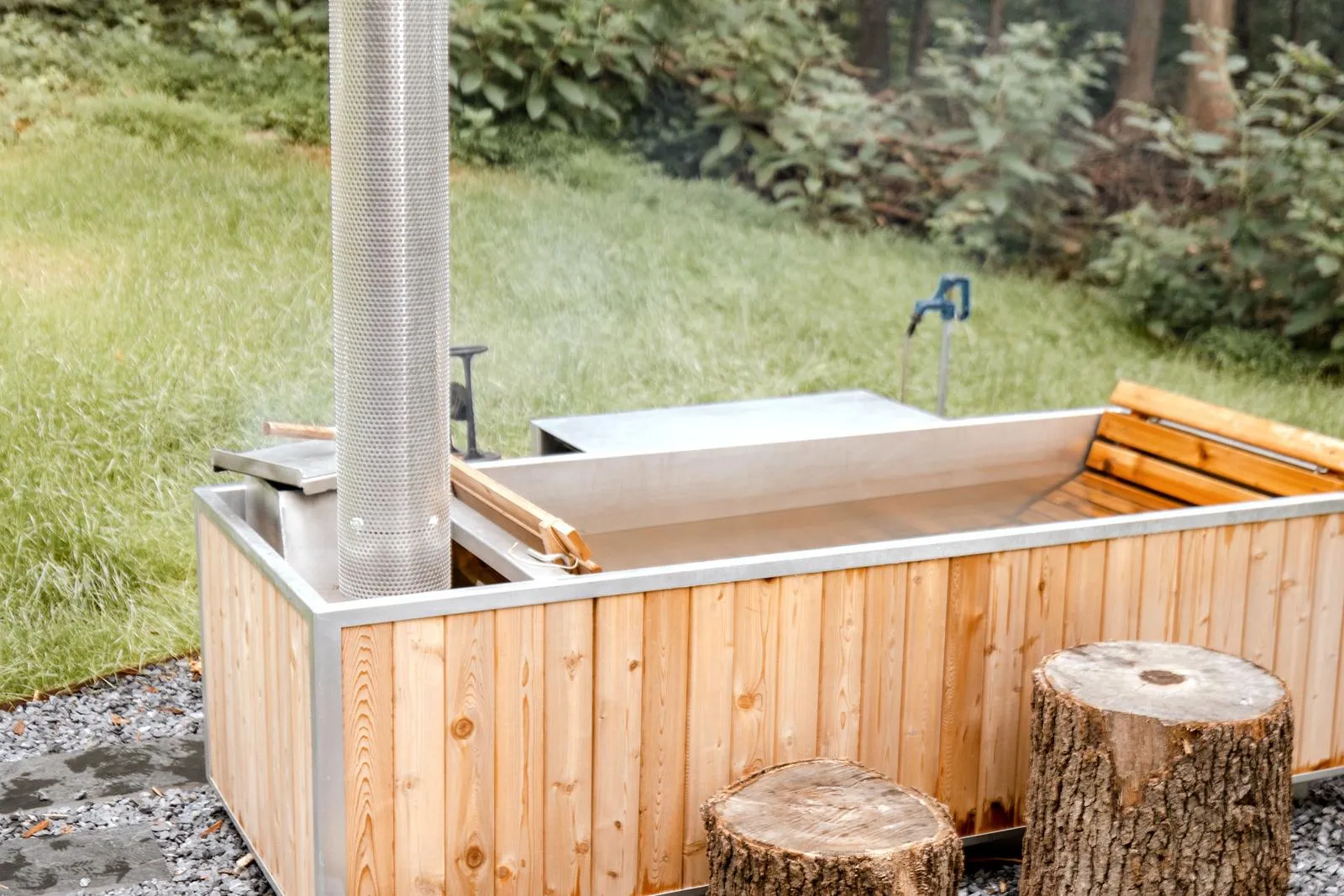 A rectangular metal tub clad in cedar, with a perforated chimney at one end and log stools on gravel in front.