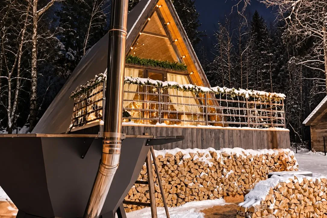 A steel hot tub with a steel chimney beside an A-frame cabin, with stacked firewood and snow around it at night.