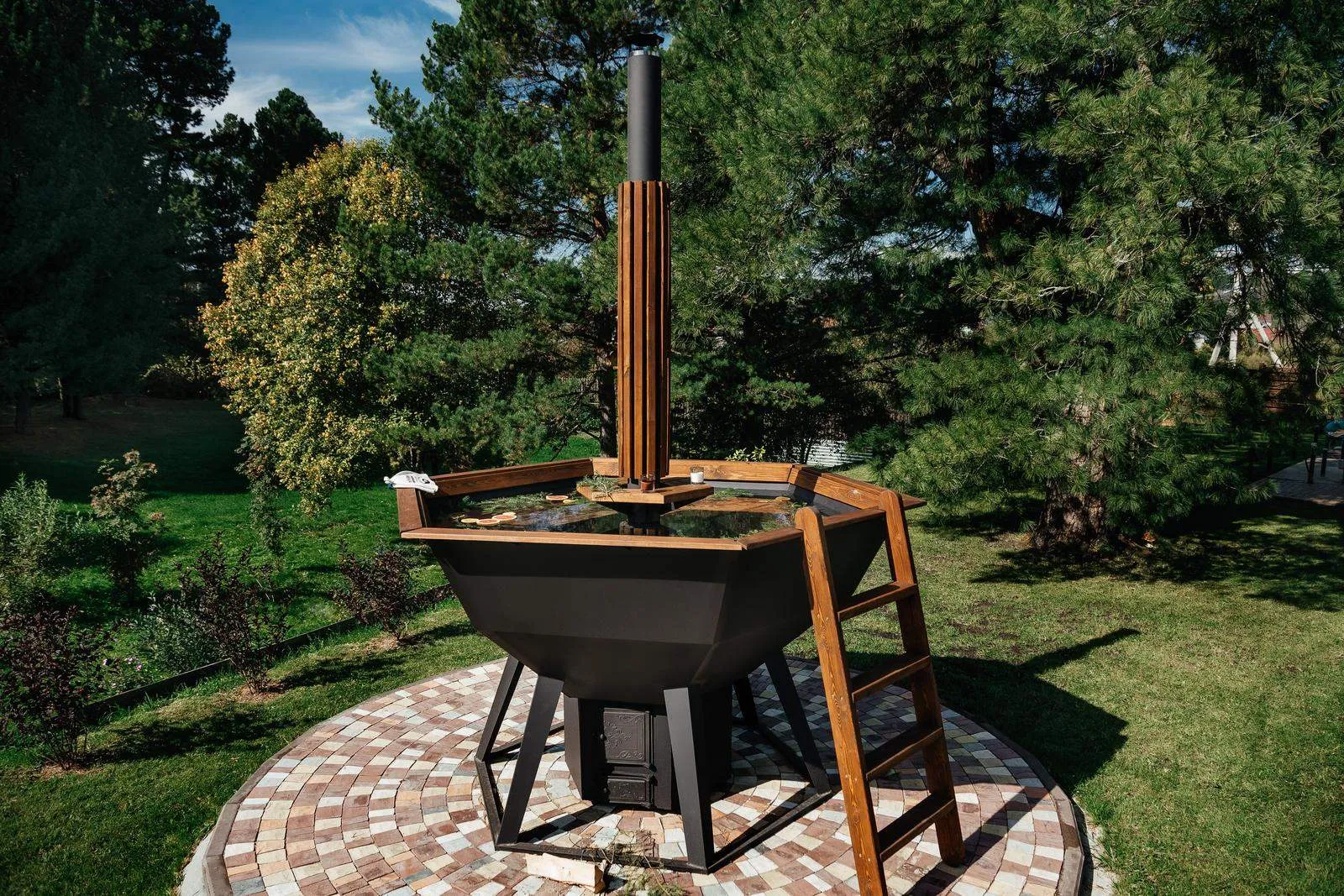 A steel hot tub with a cedar chimney guard and a wooden ladder, standing on a paved circle in a garden.