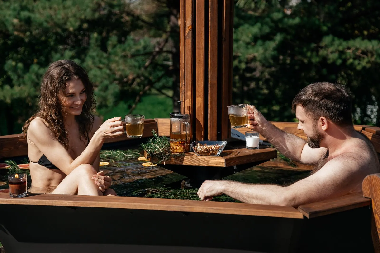 Two people share tea in a wood-fired hot tub among pine trees on a sunny day.