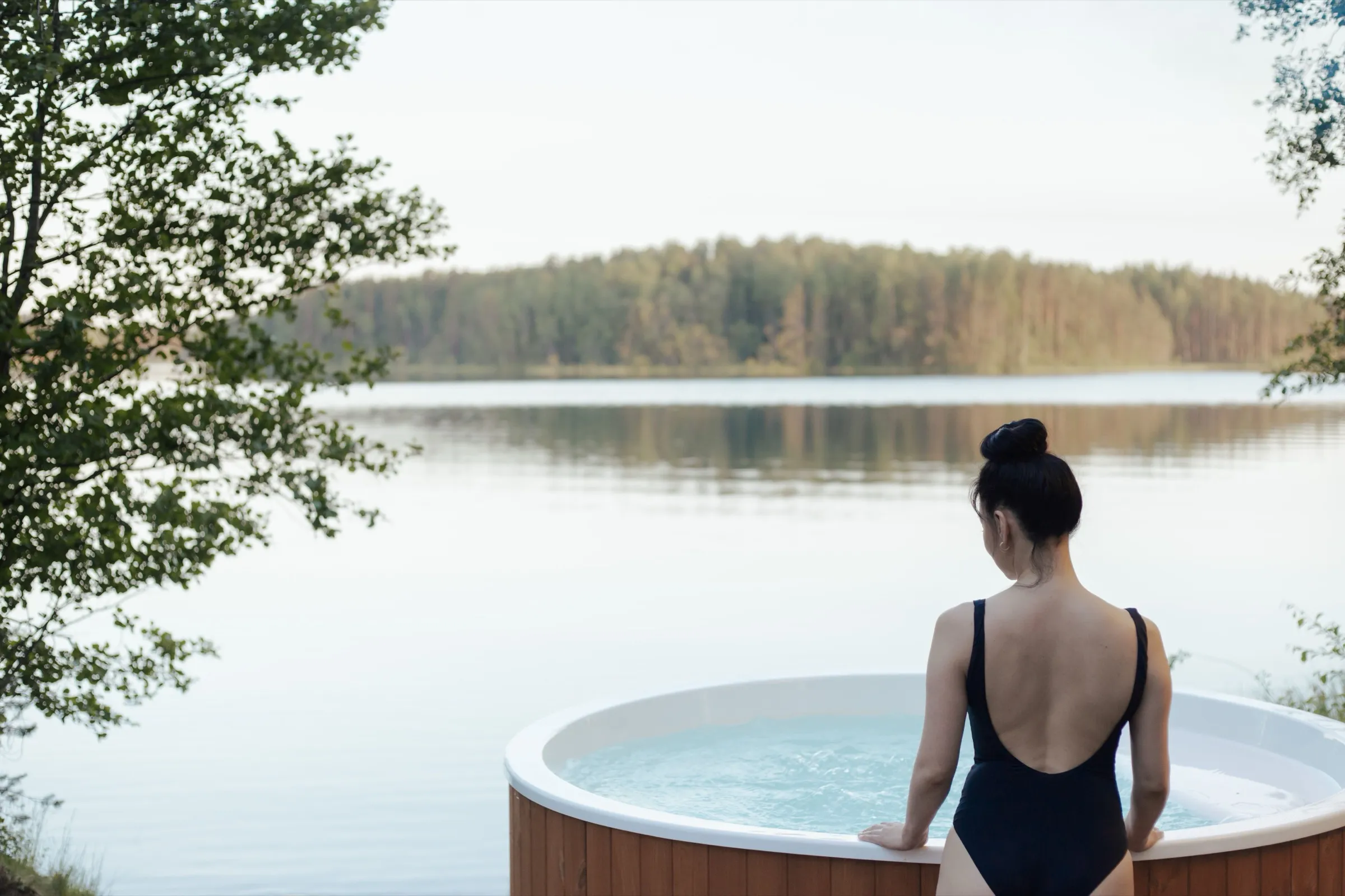 A woman in a swimsuit sits on the edge of a round hot tub, looking out over a still lake toward a treeline at dusk.
