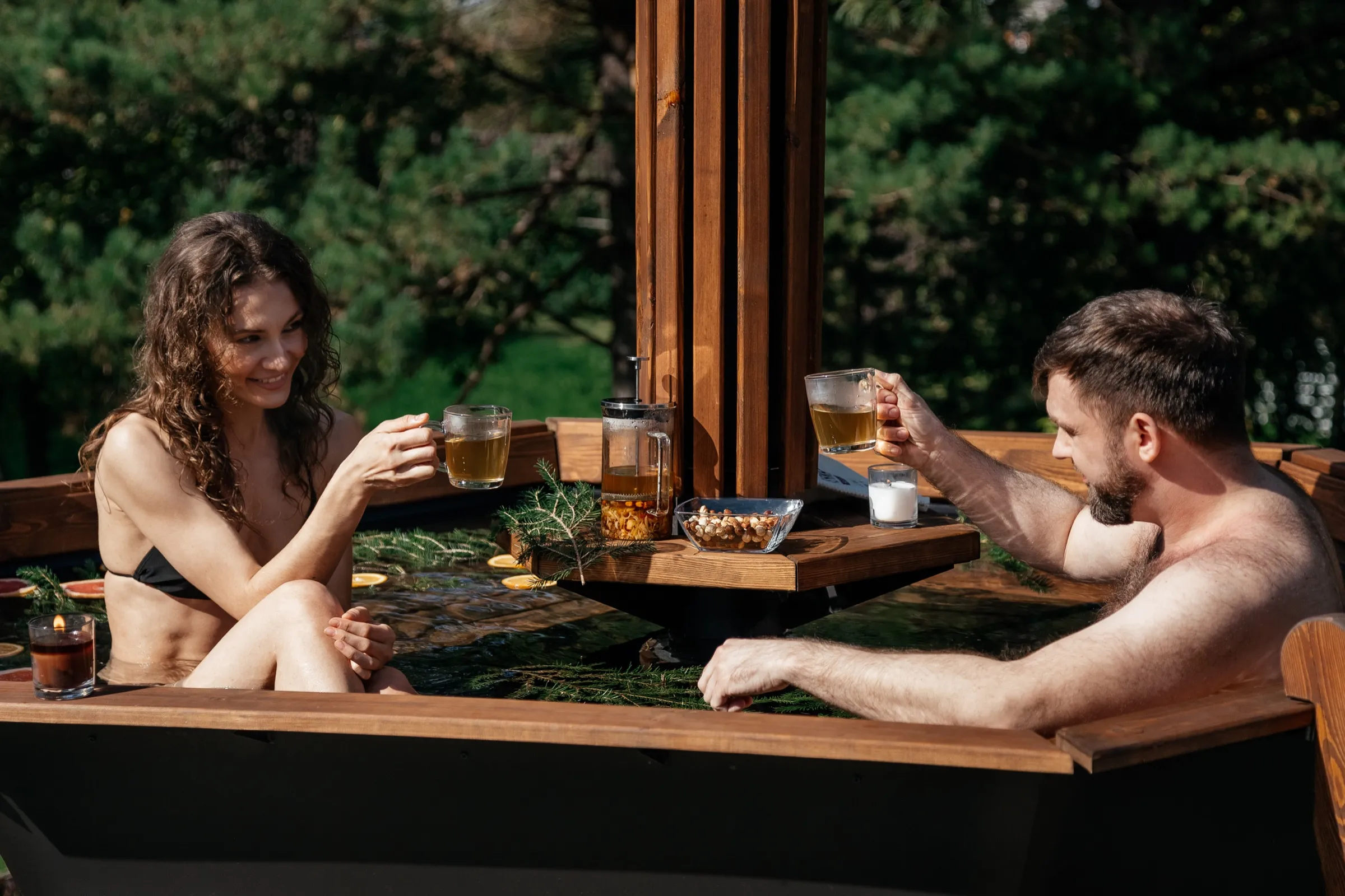 A couple sits in a wood-fired hot tub sharing tea from a French press set on the tub’s attachable table, orange slices floating in the water.